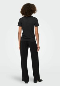 Person med krøllet hår står med ryggen til iført en ensfarvet sort t-shirt med korte ærmer, løse sorte bukser og sorte hæle.