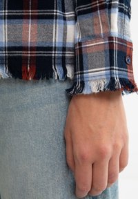 Supražinėjamas šiltas flanelinis marškiniai su mėlynos, raudonos ir baltos spalvomis; užsegami rankogaliai; dėvimi su šviesiai mėlynu džinsiniu audiniu, artimas rankovės vaizdas.