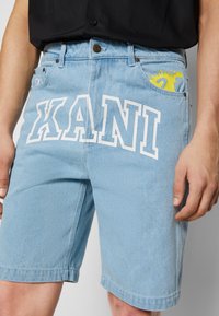 Jasnoniebieskie szorty dżinsowe z białym napisem "XANI" z przodu, z żółtym akcentem wystającym z bocznej kieszeni.