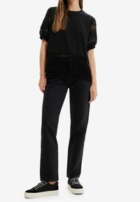 Černé tričko s průhlednými krajkovými rukávy a třásněmi na spodním lemu, zkombinované s černými džíny s vysokým pasem a černými teniskami s bílou podrážkou.