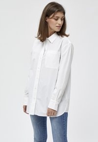 Camicia bianca a maniche lunghe con bottoni, realizzata in cotone, con taschino sul petto, colletto a punta e una vestibilità comoda, abbinata a jeans blu.