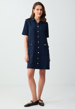 Kobieta ubrana w granatową sukienkę zapinaną na guziki z krótkimi rękawami, przednimi kieszeniami i czarnymi płaskimi butami, stojąca na jasnym, jednolitym tle.