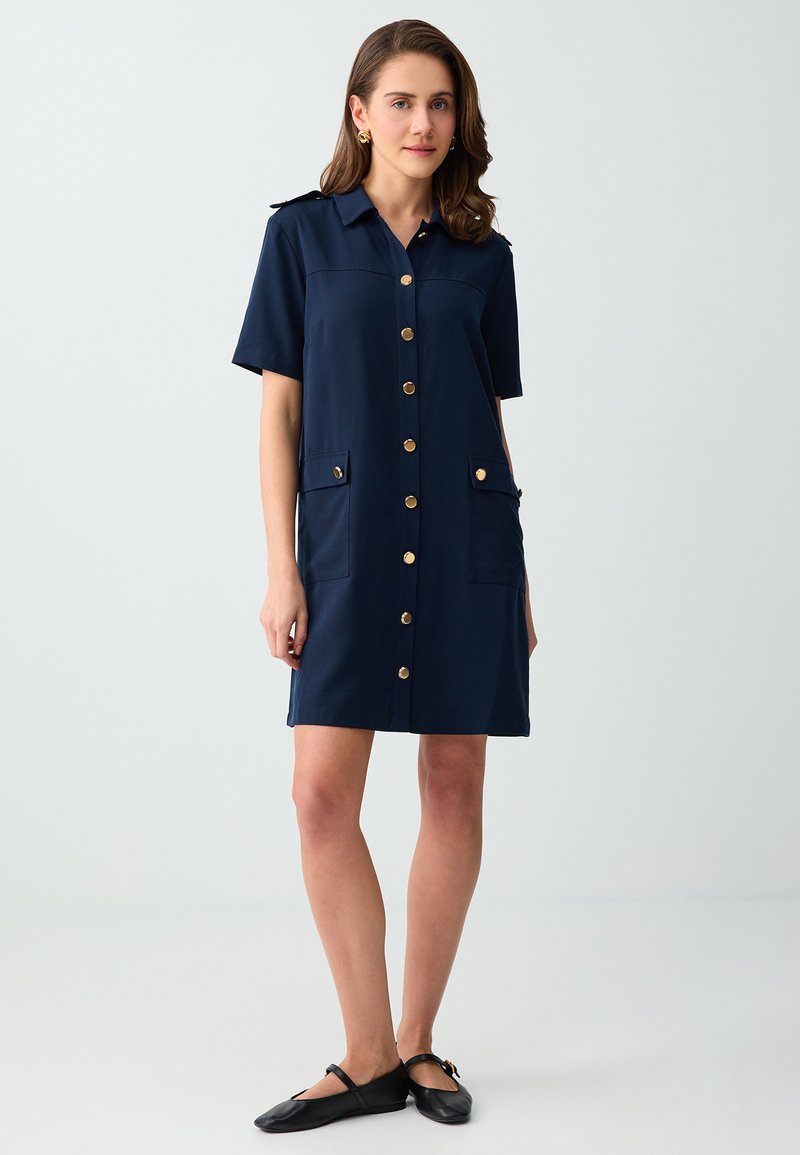 Femme portant une robe bleu marine à boutons, manches courtes, poches devant et chaussures plates noires, debout devant un fond clair uni.