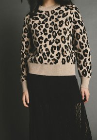 Femme portant un pull beige à motif léopard avec des poignets et un ourlet côtelés, assorti à une jupe noire texturée sur un fond gris foncé.