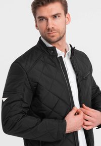 Sort quilted jakke med ståkrave, lynlås foran og lynlås lomme på brystet. Har diskrete hardwaredetaljer og en glat tekstur.