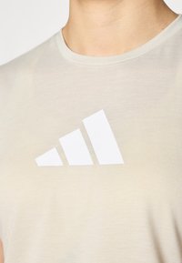 Beige kortermet t-skjorte laget av mykt stoff, med et fremtredende hvitt tre-stripes logo-design foran.