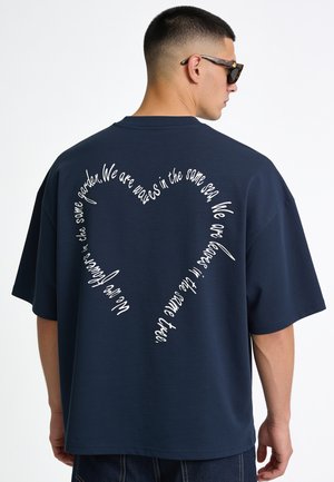 Uomo che indossa una maglietta blu navy con un design testuale a forma di cuore sulla schiena che legge una poesia su onde, mare, foglie e alberi.