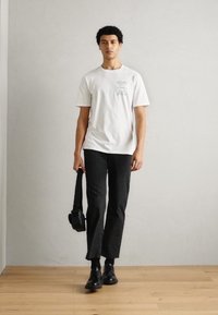 Bílý bavlněný tričko s zelenou grafikou, černé pruhované kalhoty a černé boty. Drží černou křížovou tašku. Jednoduchý, moderní outfit.