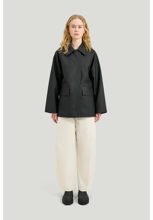 Frau steht und trägt eine schwarze übergroße Jacke, lockere weiße Hose und schwarze Stiefel vor einem schlichten hellgrauen Hintergrund.