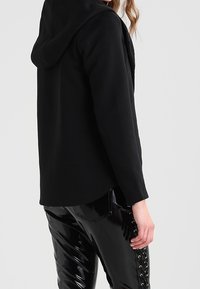 Sorte hættetrøje lavet af blødt materiale, med en afslappet pasform. Sammen med skinnende sorte bukser, der fremhæver en glat, blank tekstur.