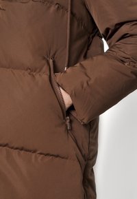 Jachetă puf dintr-un material maro, cu o textură netedă, dotată cu un buzunar lateral cu fermoar și o glugă cu șnur, evidențiind un design funcțional.