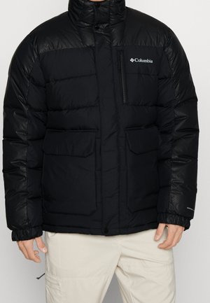 Veste isolante noire Columbia avec col haut, poche zippée sur la poitrine, deux poches avant à rabat et poignets élastiques, portée avec un pantalon beige clair.