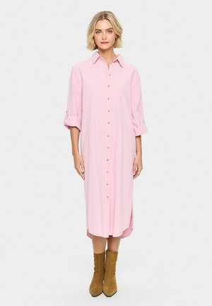 Abito camicia rosa chiaro a maniche lunghe con fronte a bottoni, maniche arrotolabili, fessure laterali, indossato con stivali alla caviglia color beige. Tessuto dal finish liscio.