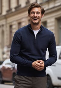 Granatowy sweter z miękkiej dzianiny, z kołnierzem na ćwierć zamka błyskawicznego i ściągaczami przy rękawach. Noszony na białym T-shircie, w połączeniu z szarymi spodniami.