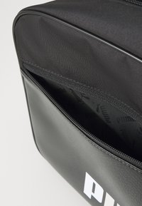 Borsa PUMA nera con apertura a zip. Realizzata in materiale sintetico, presenta una superficie liscia e un interno decorato con il nome del marchio.