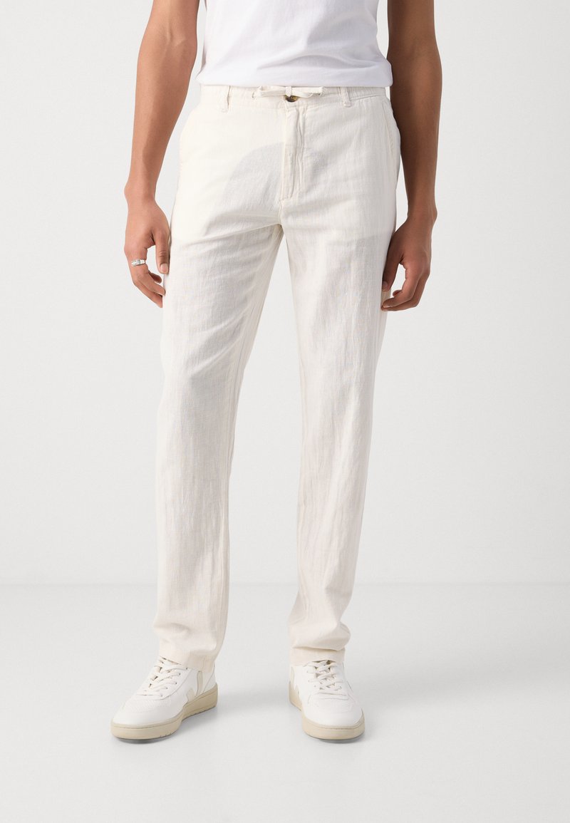 Pantalones de lino ligeros de color crema con un corte relajado, que cuentan con un cierre frontal de botón y bolsillos laterales, combinados con zapatillas blancas.