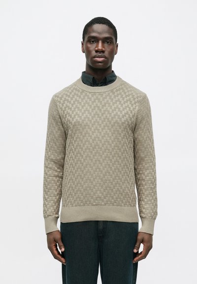 Homme portant un pull texturé beige sur une chemise noire à col et un pantalon sombre, se tenant devant un fond blanc uni.