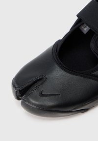 Juodas odinis batelis su apvalia nosimi ir padalinta forma. Yra juodas elastingas dirželis ir tekstūruotas paviršius. Šone yra Nike logotipas.