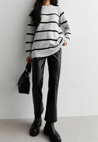 Kobieta w szarym swetrze w czarne paski, czarnych skórzanych spodniach, czarnych masywnych botkach, trzymająca czarną torebkę, stojąca przy białej ścianie.