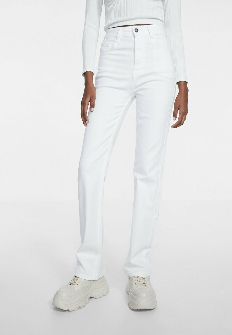 Femme portant un jean blanc taille haute à jambes droites, des baskets blanches épaisses et un crop top blanc côtelé à manches longues.