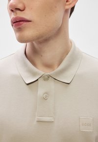 Bež polo majica iz teksturirane tkanine, s klasičnim ovratnikom, dvema gumboma in majhnim kvadratnim logotipom, izvezenim na prsih.