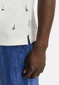 Hvid polo skjorte med farverig sejlbåd broderi, parret med krøllede blå linned shorts. Har et sideskilt med "GANT."