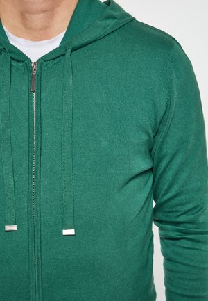 Grøn zip-up hoodie med tekstureret stof, snoretræk i hætten, sølv lynlås og metal lynlås træk. Har lange ærmer og en tætsiddende pasform.