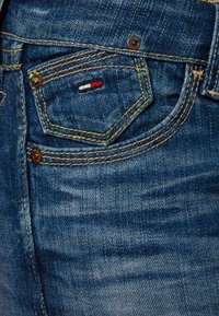 Tmavě modré džíny z denimu s přední kapsou a žlutým prošíváním. Na kapse je vyšitý malý logo v červené, bílé a modré barvě.