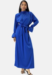 Lungo vestito di raso blu con collo alto, maniche a sbuffo e cintura annodata in vita. Tessuto liscio e silhouette fluente con una lunghezza sobria.