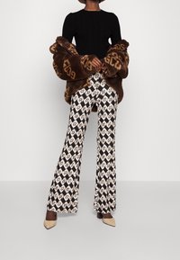 Brązowa kurtka z futra syntetycznego z oversize'owymi rękawami, czarny żebrowany top, wzorzyste spodnie wide-leg w czarnym i kremowym kolorze oraz beżowe czółenka z naostrzonym noskiem.