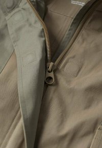 Gros plan sur une veste beige et vert olive, avec une fermeture éclair à tirette ronde, un tissu robuste et des détails en patchs carrés sur la surface.