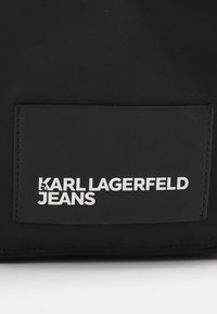 Juodas nailono krepšys su stačiakampiu pavyzdžiu, kuriame baltais rašmenimis užrašyta: "KARL LAGERFELD JEANS". Lygus paviršius su toniniais siuvimo detalėmis.