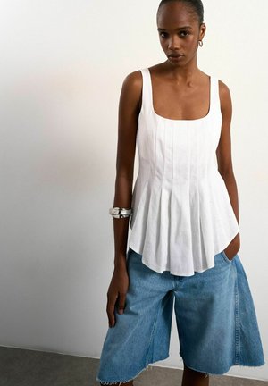 Donna che indossa un top bianco senza maniche plissettato e pantaloncini di jeans larghi fino al ginocchio, in piedi contro un muro chiaro e semplice.