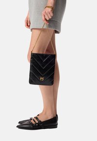 Borsa in pelle nera con motivo a zigzag, tracolla in catena dorata e un accento con il logo. Indossata con una gonna grigia e scarpe nere.