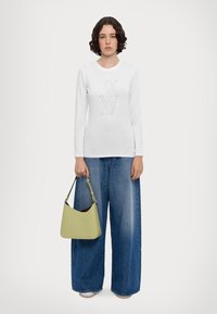 Camicia bianca a maniche lunghe con un design geometrico grigio, abbinata a jeans blu a gamba larga e una morbida borsa in pelle verde con una silhouette curvata.