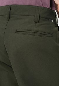 Pantalones de algodón verde con una textura suave, que cuentan con un bolsillo trasero y una etiqueta que dice "OBEY" en la cinturilla.