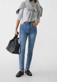 Jeans bleus taille haute avec une jambe droite, associés à un pull côtelé gris clair et des mocassins en cuir noirs ; tenant un sac noir.
