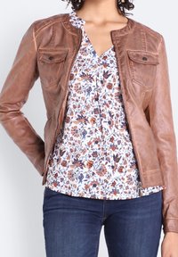 Kvinna som har på sig en brun läderjacka över en vit blommig blus med blåa och orange blommor, tillsammans med mörkblå jeans.