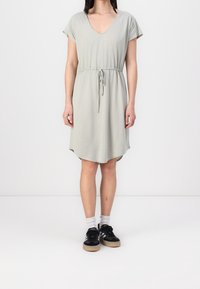Hellgraues kurzes Kleid mit kurzen Ärmeln, V-Ausschnitt, tailliertem Bund und abgerundetem Saum. Getragen mit schwarzen Turnschuhen und weißen Socken. Glatte Stoffoberfläche.