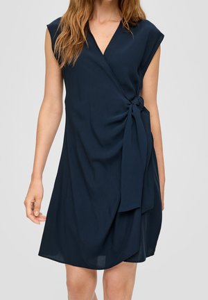 Femme portant une robe portefeuille sans manches bleu marine avec un décolleté en V profond et un détail de nœud sur le côté, debout devant un fond clair uni.