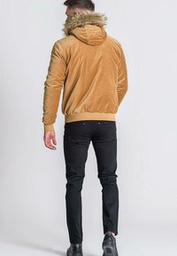 Hoodie de terciopelo marrón con capucha de piel sintética, puños y dobladillo elásticos. Combinado con jeans negros ajustados y zapatos negros, visto desde la parte trasera.