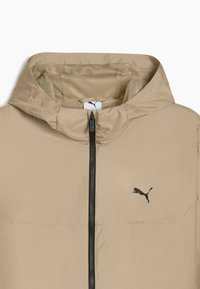 Lys beige hættejakke lavet af glat materiale, med lynlås foran, mesh-foring og et sort Puma-logo på venstre bryst.
