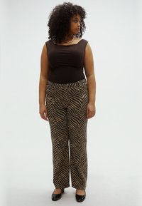 Top marrone senza maniche abbinato a pantaloni a gamba larga con motivo zebrato nero e marrone chiaro. Donna in piedi su uno sfondo semplice.