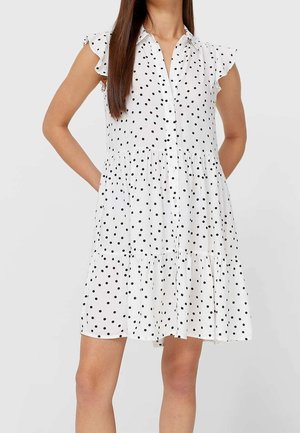 Weißes ärmelloses Kleid mit schwarzen Tupfen, vorne mit Knöpfen, Rüschen an den Schultern und gestufter Rock, getragen von einer stehenden Person.