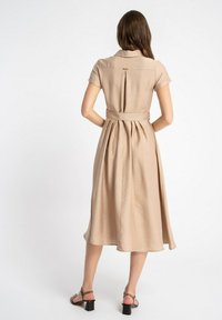 Beżowa sukienka koszulowa z krótkimi rękawami, paskiem w talii i zwiewnym dołem. Posiada plisę z tyłu i gładką fakturę. Noszona z sandałkami na paski.