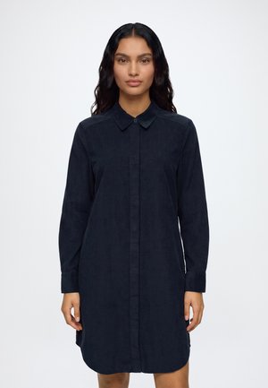 Vrouw met lang donker haar die een effen donkerblauwe overhemdjurk met lange mouwen draagt, staand tegen een witte achtergrond.