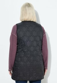 Mustat tikkivästillä varustettu liivi, jossa on aalto-ompelukuvio, korkea kaulus ja hihaton muotoilu, pukeutuneena vaaleanpunaisen pitkähihasen paidan päälle.
