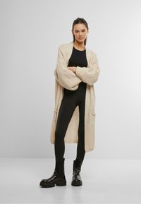 Beige Strickjacke mit lockerer Passform, gerippter Textur und Vordertaschen, getragen über einem schwarzen Oberteil und Leggings, kombiniert mit klobigen schwarzen Stiefeln.