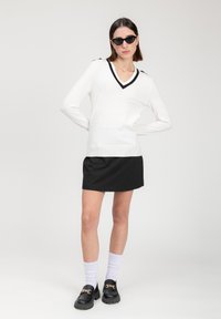 Witte V-halstrui met zwarte biezen, gecombineerd met een zwarte mini-jurk. Zwarte loafers met een kettingaccent en witte crew-sokken maken de look af.