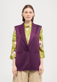 Ermeløs lila vest med en glatt tekstur og en enkelt svart knapp, lagd over en grønn blomstermønstret skjorte og beige bukser.